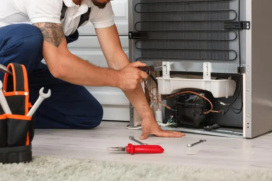 Technician diagnosing common GE refrigerator problems near the compressor and rear components