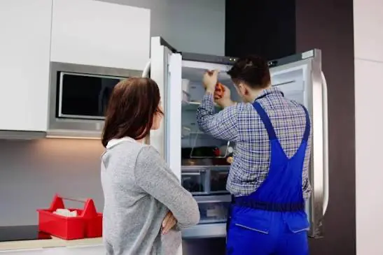 Technician inspecting a refrigerator during routine maintenance with a homeowner nearby